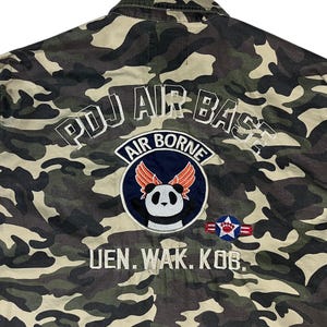May include: Camouflage jacket with "PDJ AIR BASE" and "AIR BORNE" text. It has a panda with wings patch and a military emblem. The jacket's colours include green, brown, and black.