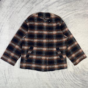 Pode incluir: Uma jaqueta de manga comprida com um padrão xadrez texturizado em tons de preto, castanho e creme. A jaqueta tem um decote redondo sem colarinho, um fecho de botão único visível no topo e dois bolsos frontais inclinados. A etiqueta Pendleton é visível.