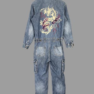 May include: A blue denim jumpsuit with a dragon embroidery on the back. The jumpsuit has long sleeves, multiple pockets, and a button-up front. It features a belt at the waist and is made of denim fabric.