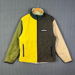 May include: A quilted jacket with a color-block design. The jacket features a combination of olive green, yellow, brown, and tan panels. It has a high collar, a zipper closure, and the "Wild Things" logo patch. The jacket is hanging on a hanger.