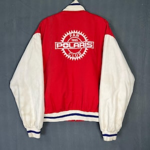 Puede incluir: Chaqueta de béisbol roja y blanca con ribete azul y blanco a rayas. La parte posterior de la chaqueta tiene un logotipo bordado en blanco que dice "Fan 1995 Polaris Club".