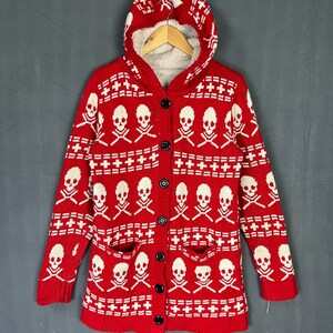 May include: Red hooded cardigan with a white skull and crossbones pattern. The sweater has a button-up front, two pockets, and a fleece-lined hood. The garment is hanging on a wooden hanger against a gray background.