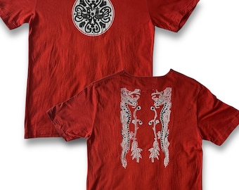 May include: Red short-sleeved t-shirt with a round, white and black ornate design on the front. The back of the shirt features a symmetrical white and black dragon-like design. The shirt is made of a soft, textured fabric.