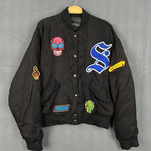 Può includere: Giacca bomber nera con diverse toppe. Le toppe includono un teschio, una nota musicale, la lettera "S" blu e la scritta "STUSSY". La giacca ha colletto, polsini e orlo a coste.