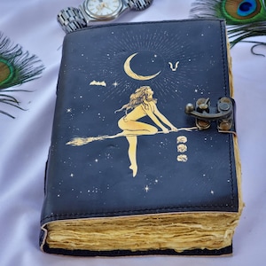 May include: A dark blue leather journal with a gold illustration of a witch riding a broom, a crescent moon, and stars. The journal has a metal clasp and aged, yellowed pages. A silver watch and peacock feather are in the background.