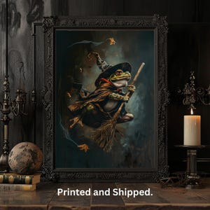 May include: Framed artwork showing a frog witch riding a broomstick. The frog is dressed in a pointed hat and cape, set against a dark background. The black frame is ornate. The artwork is displayed on a wooden surface. The image has a fantasy theme.