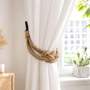May include: A natural rope curtain tieback knotted around white curtains. The tieback is made of thick, twisted rope and is secured to the wall with a black metal bracket. The curtains are hanging in front of a window with a view of greenery.