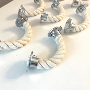May include: A set of curved rope handles with silver metal mounting plates. The handles are made of thick, off-white rope, and the metal plates have a shiny finish. The handles are arranged on a white surface, suitable for a nautical theme.