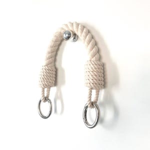 May include: A cream-colored rope handle with silver metal rings and a central mounting point. The rope is thick and tightly wound, creating a textured appearance. The handle is designed for hanging or decorative purposes.