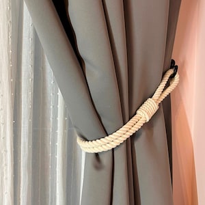 May include: A white rope curtain tieback with a black metal hook. The tieback is holding back a gray curtain.