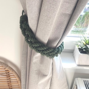 May include: A green rope curtain tieback holds a beige curtain. The braided tieback is made of thick, textured rope. A houseplant in a white pot sits on a windowsill in the background.