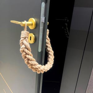 Könnte beinhalten: Ein goldener Türgriff mit einem Türstopper aus Seil. Der Türstopper ist aus Naturseil gefertigt und um den Griff gewickelt.