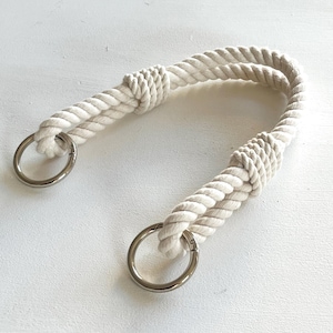 Könnte beinhalten: Cremefarbener Seilgriff mit zwei silbernen Metallringen. Das Seil ist dick und gedreht, mit Knoten, die die Ringe sichern. Der Griff ist gebogen, was darauf hindeutet, dass er zum Tragen oder Aufhängen von Gegenständen bestimmt ist. Der Hintergrund ist eine weiße Oberfläche.