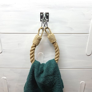 Puede incluir: Un toallero de temática náutica hecho de cuerda gruesa y trenzada, suspendido de un soporte metálico. Una toalla verde azulado doblada cuelga del anillo. El fondo es una pared de madera pintada de blanco con detalles decorativos.