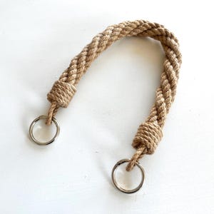 May include: A natural-toned rope handle with two silver-colored metal rings. The rope is tightly braided and knotted at each end, creating a sturdy and decorative accessory. The handle is designed for use with bags or other items.