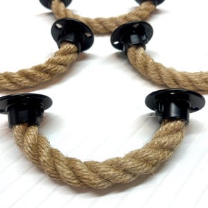 Op de afbeelding: Vier zwarte metalen touwgrepen met natuurlijk jute touw. Het touw is gedraaid en heeft een natuurlijke bruine kleur. De grepen zijn ontworpen voor gebruik op kasten, lades of deuren.