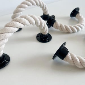 Puede incluir: Varias manijas de cuerda con placas de montaje negras están dispuestas sobre una superficie blanca. Las manijas están hechas de cuerda gruesa, blanquecina, curvada en semicírculo. Las placas negras tienen orificios para tornillos.