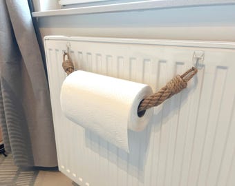 May include: A white paper towel roll suspended by a thick, brown rope. The rope is attached to the radiator with clear adhesive hooks. The paper towel roll is centered in the image, with a portion of the paper unfurled.