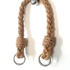 May include: Braided jute rope handles with silver-colored metal rings. The rope is a natural tan color, and the handles are knotted at the ends. The handles are attached to a silver-colored metal hook.