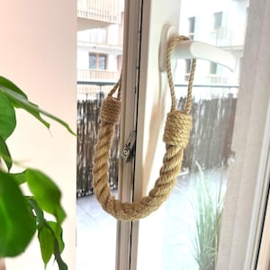 May include: A beige rope doorstop with a loop at the top, hanging on a white door handle. The doorstop is made of natural fibre rope.