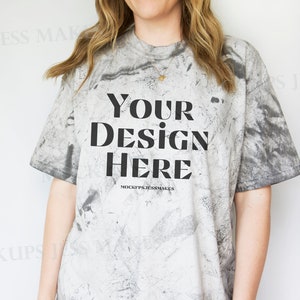 Puede incluir: Una camiseta blanca con un patrón de tie-dye negro y gris. La camiseta tiene el texto "YOUR DESIGN HERE" impreso en la parte delantera. El texto "MOCKUPSJESSMAKES" está impreso debajo del diseño.