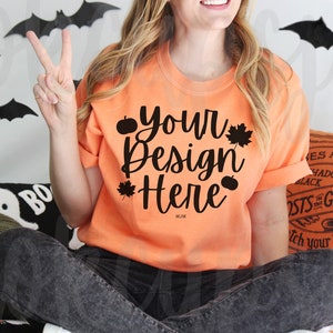 May include: A bright orange t-shirt with a black design that says "your design here" with pumpkins and leaves. The shirt is being modeled by a person sitting down.