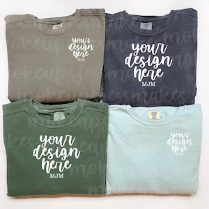 May include: Four folded sweatshirts in different colors: olive green, dark green, dark gray, and light blue. Each sweatshirt has a white text design that says "your design here" and "MJM".