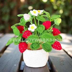 May include: A crocheted strawberry plant in a white pot. The plant features bright red strawberries, green leaves, white flowers, and the text "PINK LADY WORKSHOP". The pot is sitting on a wooden surface.