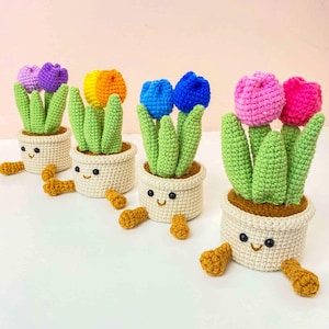 May include: Crocheted tulip plants in white pots with smiling faces. The tulips are in various colors: purple, yellow, orange, blue, and pink. Green leaves and brown soil complete the look. Several small, brown crocheted pieces are scattered around the pots.