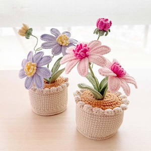 May include: Two crocheted flower arrangements in pots. The flowers are pink, purple, and white. The pots are beige with a brown rim.