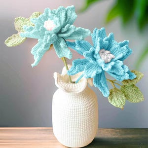 May include: A white crocheted vase displays two handmade, light blue crocheted flowers with pink centres and green leaves. The flowers are the focal point, with the vase providing a simple, textured base. The arrangement is a charming, handcrafted decorative item.