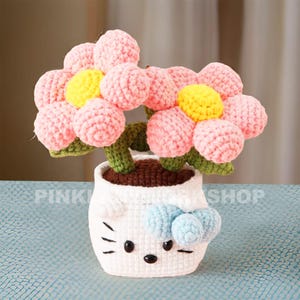 May include: A crocheted flower arrangement in a white pot. The flowers are pink with yellow centers, green stems, and leaves. The pot is white with a cartoon face and a blue bow. The text "PINK SHOP" is visible on the pot.