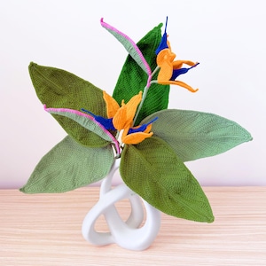 Op de afbeelding: Een handgemaakt gehaakt paradijsvogelbloemstuk. Het arrangement heeft grote groene bladeren, oranje en blauwe bloemen en een witte vaas. De bloemen hebben roze en blauwe accenten, waardoor een levendig decoratief stuk ontstaat.