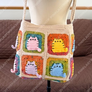 May include: A beige crochet tote bag featuring four square panels, each displaying a different colored cat motif. The cats are pink, yellow, lavender, and blue, all with black stitched details. The bag has a wide shoulder strap and a neutral border.