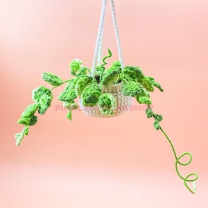 May include: A handmade crochet hanging planter with a trailing green plant. The white planter is suspended by a white rope. The plant has bright green leaves and vines, creating a whimsical, decorative piece.