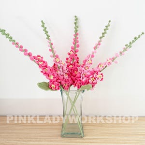 May include: A bouquet of handmade crochet flowers in a clear glass vase. The flowers are various shades of pink with yellow centers and green stems and leaves. The text "PINKLADYWORKSHOP" is at the bottom of the image.