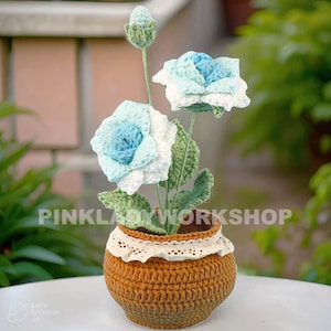 Op de afbeelding: Een handgemaakt gehaakt bloemstuk in een bruine pot. Het arrangement heeft blauwe en witte rozen met groene bladeren en een knop. De pot heeft een kanten rand en de tekst "PINKLADYWORKSHOP".