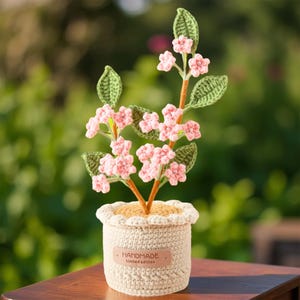 May include: A handmade crochet plant with pink flowers and green leaves in a cream-colored pot. The pot has a decorative edge and a small tag that reads "HANDMADE Limited edition". The plant is on a wooden surface.
