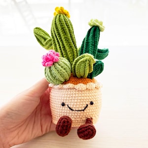 May include: A crocheted cactus plant with pink and yellow flowers in a beige pot with a smiling face and brown legs.