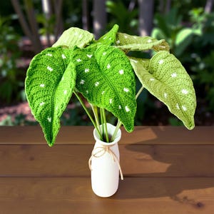 May include: A handcrafted crochet plant arrangement featuring vibrant green leaves with white polka dots. The leaves are arranged in a white vase tied with twine, creating a unique and decorative piece. The arrangement is set on a wooden surface.