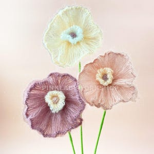 May include: Three handmade, fluffy flowers in shades of cream, peach, and mauve, with white and cream centers. The flowers have green stems and are set against a soft pink background. The flowers appear to be made of yarn or a similar fiber.