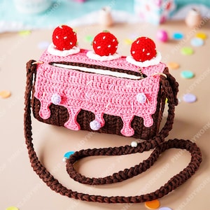 May include: A crocheted purse shaped like a cake, featuring a pink top layer with brown 'chocolate' dripping down. The cake is topped with three red strawberry decorations. The purse has a brown strap and zipper closure.