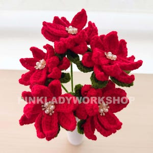 Op de afbeelding: Een boeket van handgemaakte, rode gehaakte poinsettia's met groene bladeren en parelaccenten. De bloemen staan in een witte vaas. De tekst "PINKLADYWORKSHOP" is zichtbaar.