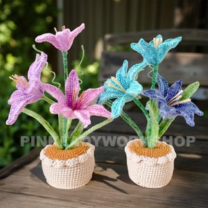 May include: Two crochet flower arrangements in cream-colored pots. One arrangement features pink and purple flowers, the other has blue and teal blooms. The flowers have green stems and are set on a wooden surface.