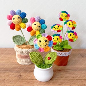 May include: Crocheted flowers with smiling faces in various colors, including red, yellow, blue, and green, are displayed in small pots. The flowers have green leaves and are arranged on a wooden surface. Some pots are crocheted, and one has a ladybug.