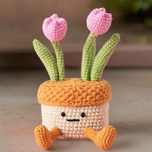 Op de afbeelding: Een gehaakte bloempot met een glimlachend gezicht en twee roze tulpen. De pot is oranje en wit met oranje poten.