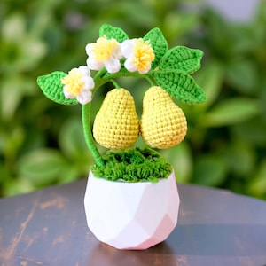 May include: A handmade crochet pear tree in a white geometric pot. The tree has two yellow pears, green leaves, and white and yellow flowers. The pot is filled with green moss, creating a charming decorative piece.