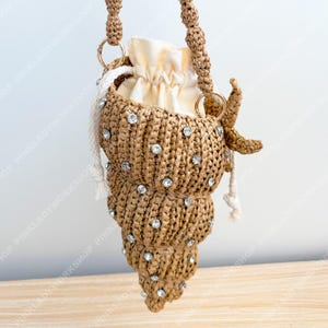 Könnte beinhalten: Handgefertigte, muschelförmige Handtasche aus gewebtem, hellbraunem Material. Die Tasche ist mit funkelnden Strasssteinen verziert und hat einen cremefarbenen Kordelzugbeutel im Inneren. Sie hat einen geflochtenen Griff und goldfarbene Ringe.