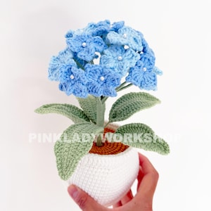 Puede incluir: Una planta de hortensia azul tejida a ganchillo en una maceta blanca. La flor tiene pétalos azul claro con detalles de perlas blancas. Las hojas y el tallo verdes contrastan con las flores azules. La maceta es blanca con base marrón.