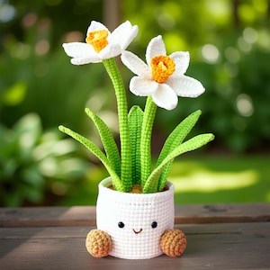 May include: A crocheted white flower pot with a smiling face, holding two white daffodils with yellow centers and green stems and leaves. The pot has two brown crocheted feet. The background is a blurred green.